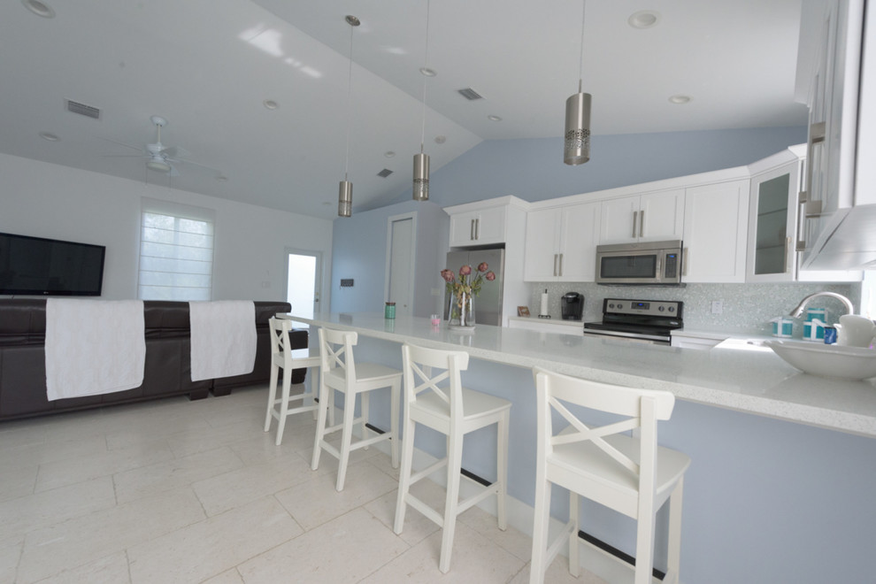 Photo of a mid-sized beach style u-shaped eat-in kitchen in Miami with a drop-in sink, shaker cabinets, white cabinets, solid surface benchtops, multi-coloured splashback, mosaic tile splashback, stainless steel appliances, ceramic floors and a peninsula.