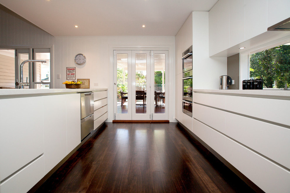 This is an example of a large contemporary galley eat-in kitchen in Brisbane with flat-panel cabinets, white cabinets, quartz benchtops, brown splashback, glass sheet splashback, stainless steel appliances, dark hardwood floors and with island.