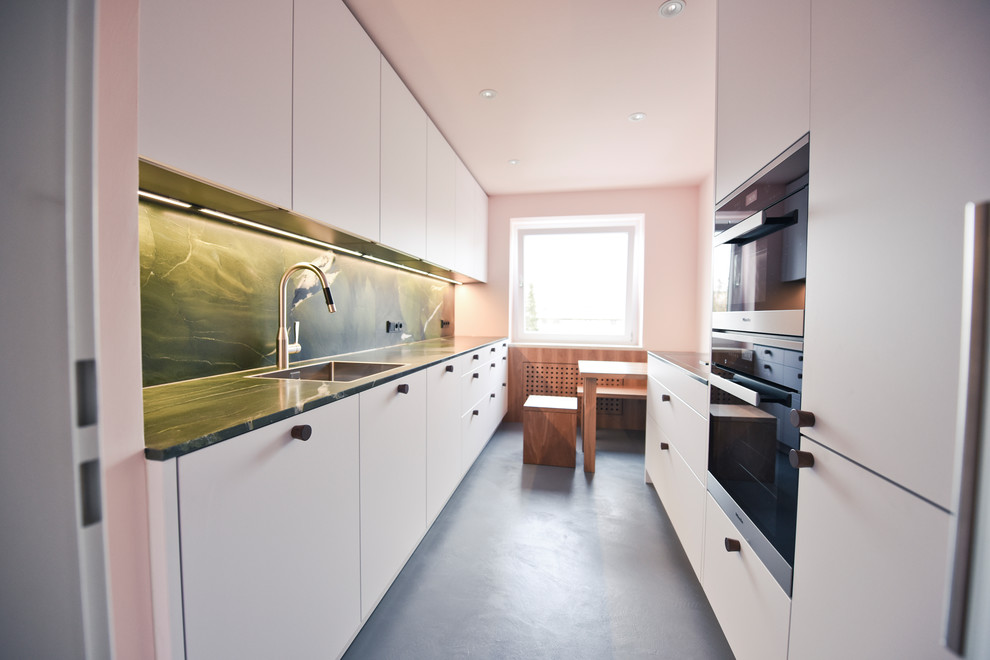 Photo of a mid-sized scandinavian galley separate kitchen in Munich with a single-bowl sink, flat-panel cabinets, white cabinets, granite benchtops, green splashback, stone slab splashback, black appliances, concrete floors, no island, grey floor and green benchtop.