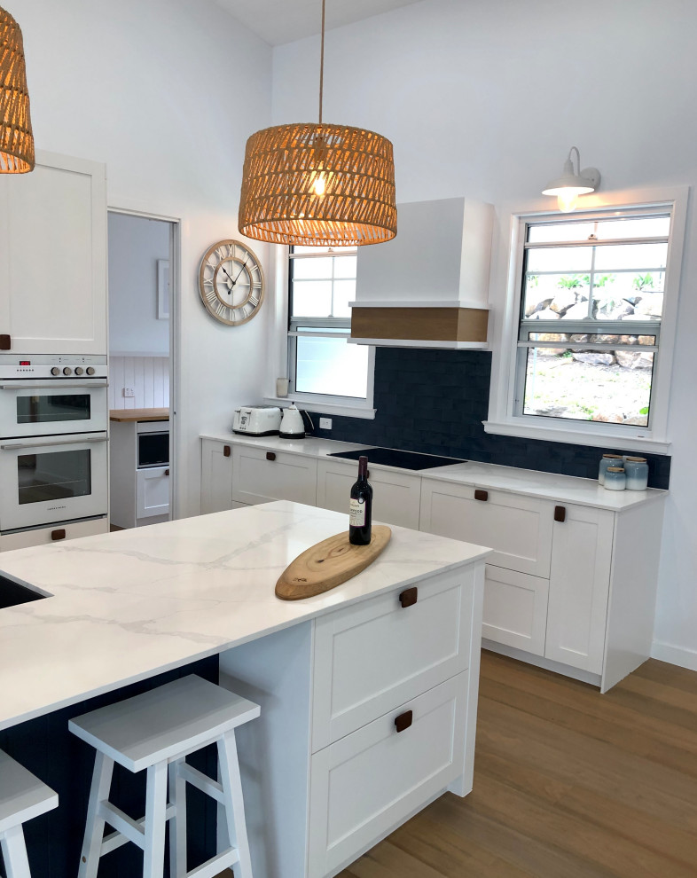This is an example of a large beach style galley open plan kitchen in Gold Coast - Tweed with a double-bowl sink, shaker cabinets, white cabinets, quartz benchtops, blue splashback, subway tile splashback, white appliances, medium hardwood floors, with island, brown floor and white benchtop.