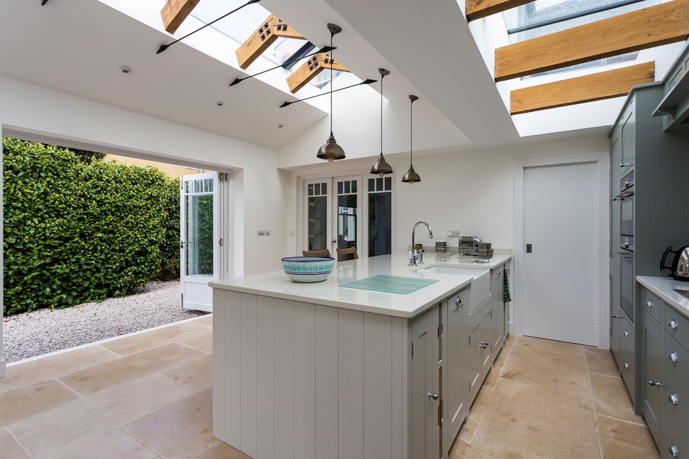 This is an example of a country kitchen in Other with a farmhouse sink, recessed-panel cabinets, green cabinets, solid surface benchtops, white splashback, ceramic splashback, stainless steel appliances, limestone floors, with island, beige floor and white benchtop.
