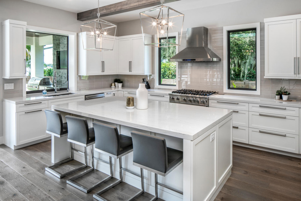 Photo of a large modern u-shaped eat-in kitchen in Tampa with a double-bowl sink, shaker cabinets, white cabinets, quartz benchtops, grey splashback, glass tile splashback, stainless steel appliances, medium hardwood floors, with island, brown floor, grey benchtop and exposed beam.