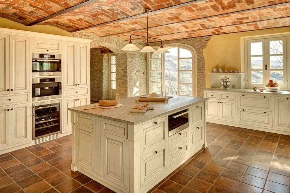 Large country l-shaped kitchen in Bologna with raised-panel cabinets, white cabinets, granite benchtops, white splashback, stainless steel appliances, terra-cotta floors, with island and brown floor.