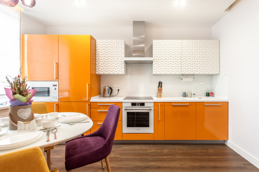 Inspiration for a small contemporary single-wall open plan kitchen in Yekaterinburg with white splashback, glass sheet splashback, white appliances, vinyl floors, brown floor, a drop-in sink, flat-panel cabinets, orange cabinets, solid surface benchtops, no island and white benchtop.