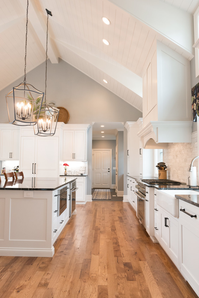 Design ideas for a large traditional eat-in kitchen in Other with a farmhouse sink, beaded inset cabinets, white cabinets, granite benchtops, marble splashback, stainless steel appliances, light hardwood floors, with island, brown floor and black benchtop.
