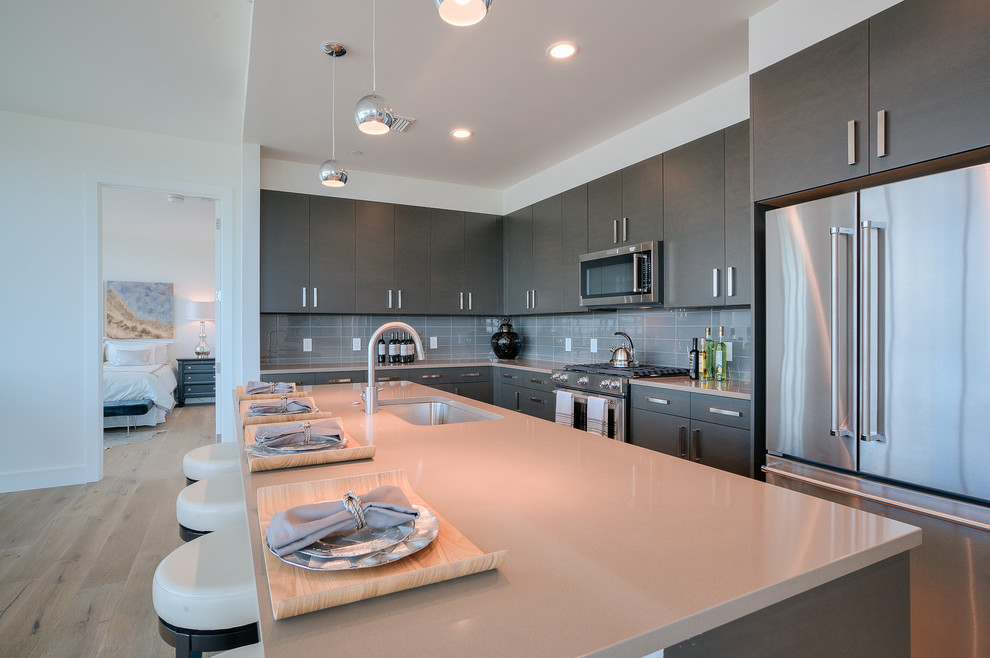 Inspiration for a large contemporary l-shaped kitchen in Austin with an undermount sink, flat-panel cabinets, grey cabinets, solid surface benchtops, grey splashback, glass tile splashback, stainless steel appliances, light hardwood floors and with island.