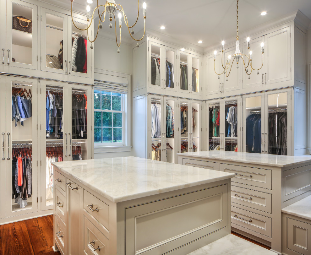 This is an example of an expansive transitional gender-neutral walk-in wardrobe in New Orleans with glass-front cabinets, white cabinets, medium hardwood floors, brown floor and vaulted.