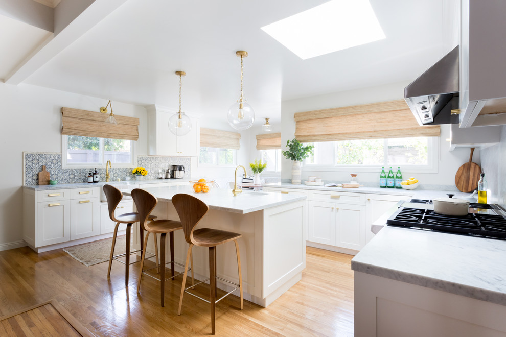 Beach style u-shaped open plan kitchen in New York with a farmhouse sink, shaker cabinets, white cabinets, marble benchtops, blue splashback, stainless steel appliances, light hardwood floors, with island and beige floor.