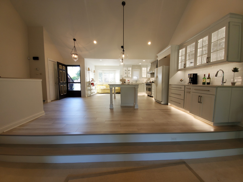 Photo of a mid-sized transitional l-shaped eat-in kitchen in Chicago with an undermount sink, glass-front cabinets, white cabinets, quartzite benchtops, white splashback, ceramic splashback, stainless steel appliances, laminate floors, with island, beige floor, white benchtop and wallpaper.