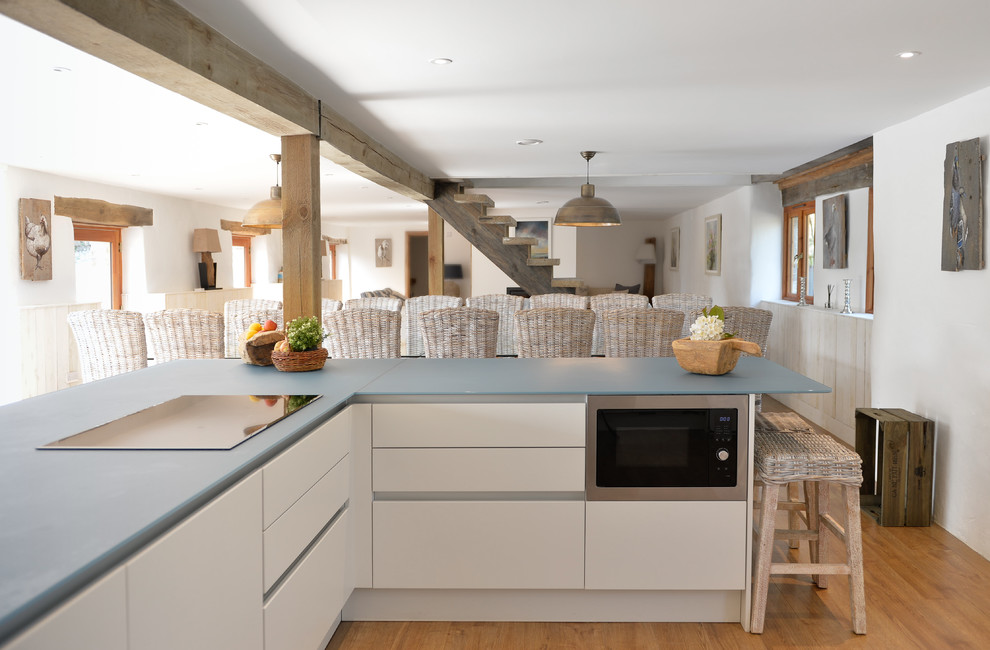 This is an example of a large country l-shaped open plan kitchen in Cornwall with a drop-in sink, flat-panel cabinets, white cabinets, glass benchtops, blue splashback, black appliances, with island and blue benchtop.