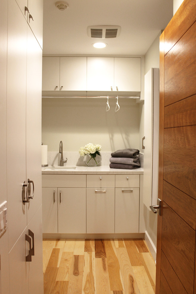 Photo of a mid-sized contemporary l-shaped dedicated laundry room in Los Angeles with an undermount sink, flat-panel cabinets, white cabinets, quartzite benchtops, grey walls, a stacked washer and dryer and light hardwood floors.