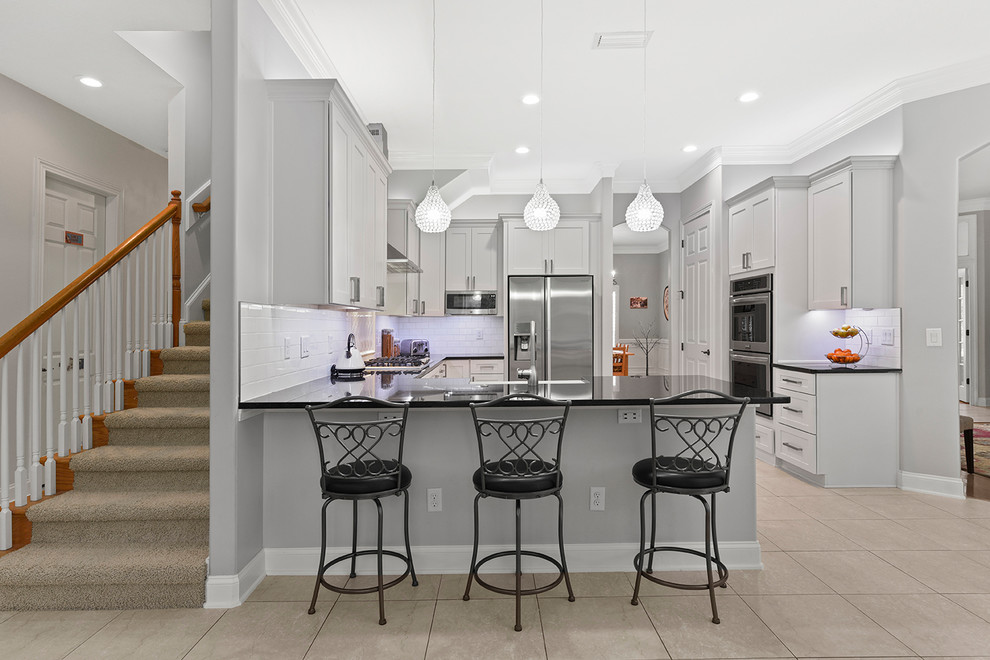 Inspiration for a mid-sized modern l-shaped eat-in kitchen in Jacksonville with a farmhouse sink, shaker cabinets, white cabinets, granite benchtops, white splashback, subway tile splashback, stainless steel appliances, ceramic floors, a peninsula, beige floor and black benchtop.