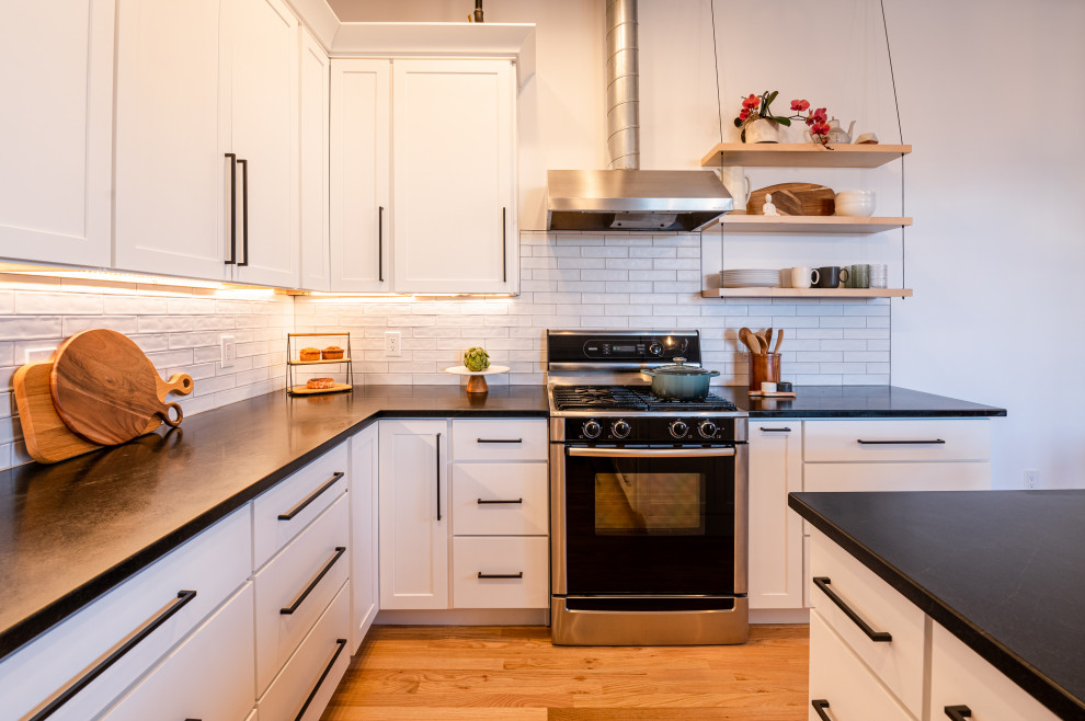 This is an example of a large industrial l-shaped eat-in kitchen in Denver with an integrated sink, shaker cabinets, white cabinets, soapstone benchtops, white splashback, ceramic splashback, stainless steel appliances, light hardwood floors, with island, black benchtop and exposed beam.