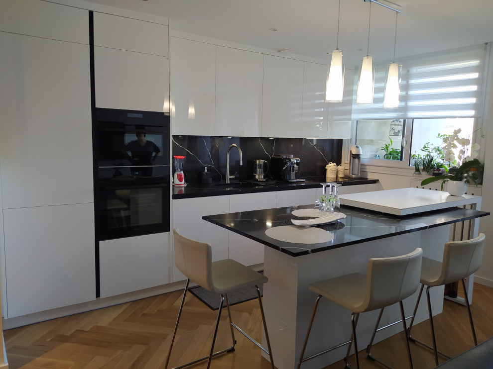 This is an example of a mid-sized modern galley open plan kitchen in Paris with an integrated sink, beaded inset cabinets, white cabinets, quartzite benchtops, black splashback, black appliances, medium hardwood floors, with island, brown floor and black benchtop.