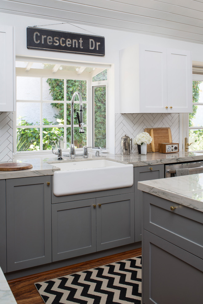 Mid-sized elegant u-shaped medium tone wood floor kitchen pantry photo in Los Angeles with a farmhouse sink, shaker cabinets, gray cabinets, quartz countertops, white backsplash, porcelain backsplash, stainless steel appliances and an island