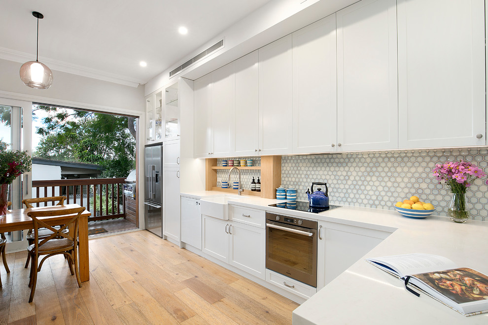 This is an example of a transitional l-shaped eat-in kitchen in Sydney with white cabinets, blue splashback, stainless steel appliances, a farmhouse sink, shaker cabinets, light hardwood floors, a peninsula, white benchtop and mosaic tile splashback.