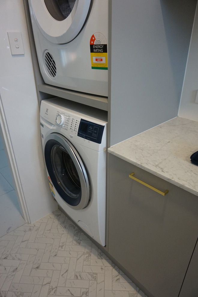 This is an example of a small contemporary l-shaped utility room in Sydney with a single-bowl sink, flat-panel cabinets, grey cabinets, quartz benchtops, white walls, porcelain floors, a stacked washer and dryer, multi-coloured floor and multi-coloured benchtop.