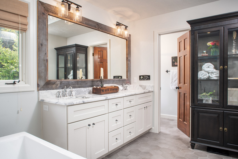 This is an example of a mid-sized country master bathroom in Omaha with shaker cabinets, white cabinets, a freestanding tub, white walls, porcelain floors, an undermount sink, marble benchtops, grey floor and white benchtops.