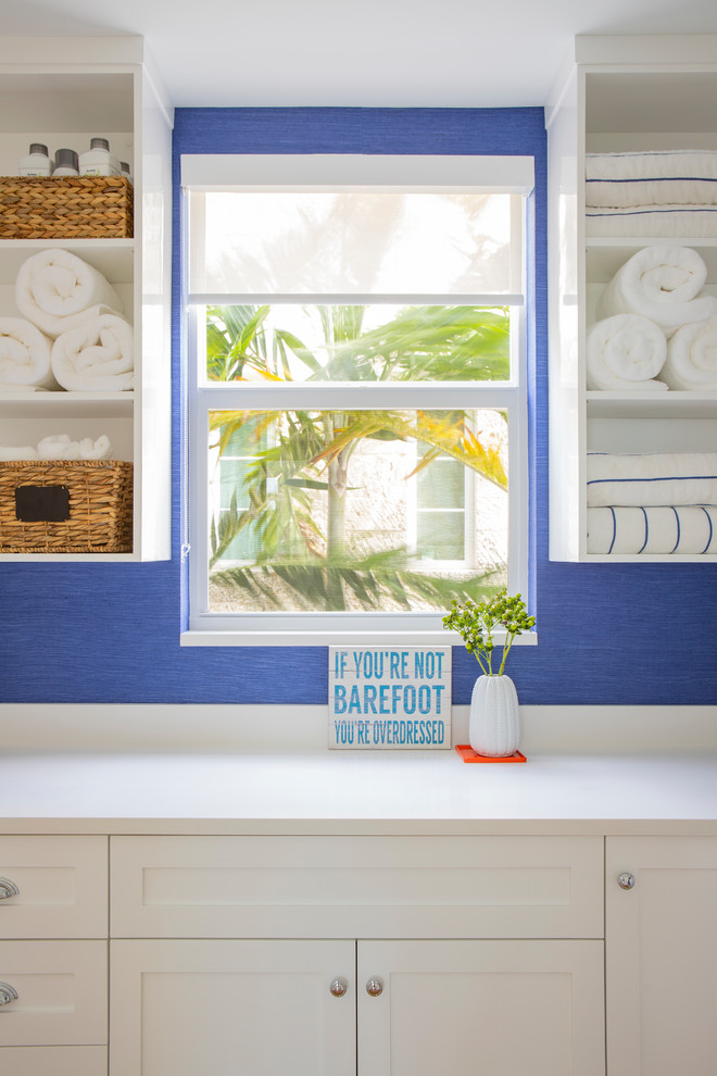Design ideas for a mid-sized beach style galley dedicated laundry room in Miami with an undermount sink, shaker cabinets, white cabinets, quartzite benchtops, blue walls, light hardwood floors, a stacked washer and dryer, beige floor and white benchtop.