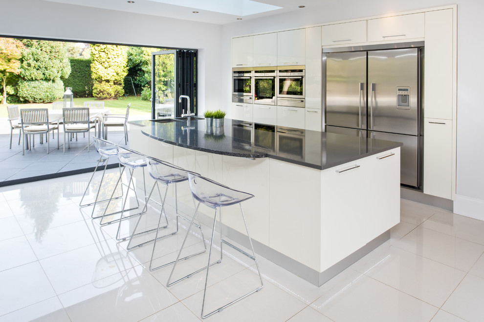 Photo of a large modern single-wall open plan kitchen in West Midlands with a double-bowl sink, beige cabinets, quartzite benchtops, stainless steel appliances, a peninsula, beige floor and black benchtop.