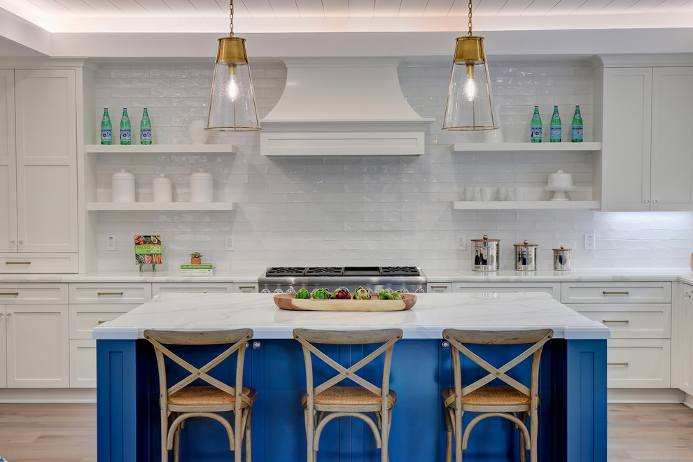 Example of a beach style l-shaped light wood floor and beige floor kitchen design in Los Angeles with shaker cabinets, white cabinets, marble countertops, white backsplash, subway tile backsplash, stainless steel appliances, an island and white countertops
