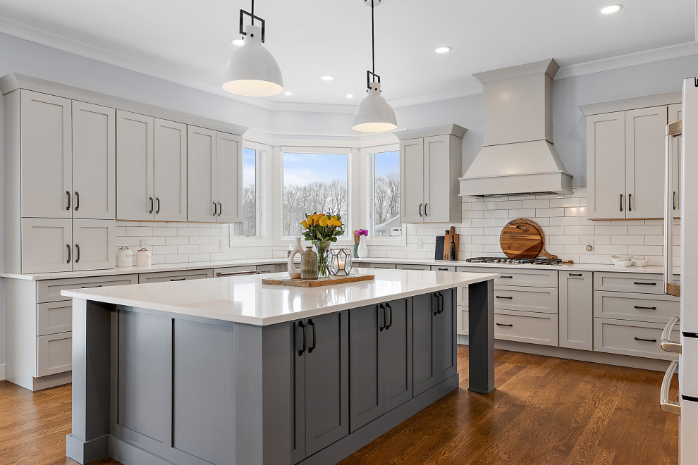 This is an example of a large transitional u-shaped eat-in kitchen in Detroit with an undermount sink, shaker cabinets, white cabinets, quartz benchtops, white splashback, ceramic splashback, white appliances, medium hardwood floors, with island, brown floor, white benchtop and vaulted.