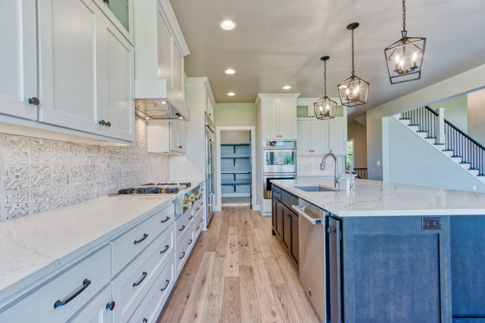 Example of a large transitional l-shaped light wood floor and brown floor eat-in kitchen design with an undermount sink, shaker cabinets, white cabinets, quartz countertops, white backsplash, ceramic backsplash, stainless steel appliances, an island and white countertops