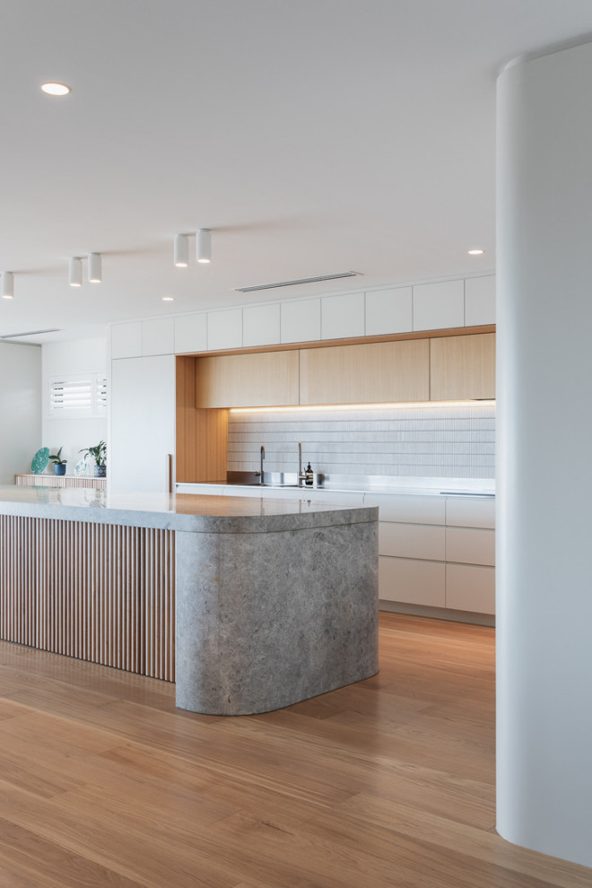 Photo of a modern galley kitchen in Perth with flat-panel cabinets, white cabinets, white splashback, stainless steel appliances, medium hardwood floors, with island, brown floor and grey benchtop.
