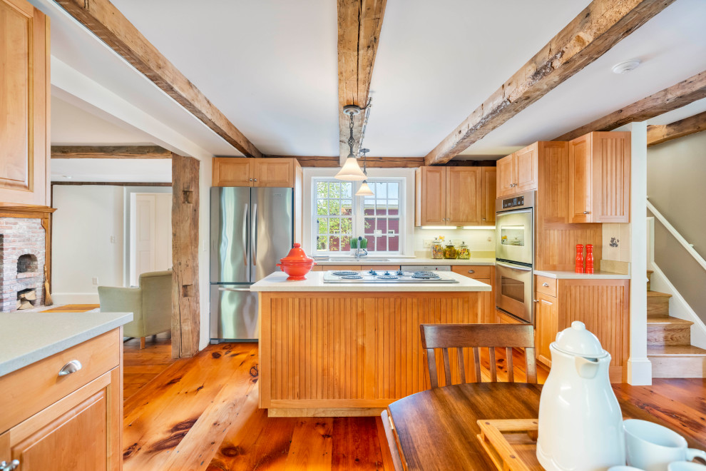This is an example of a country l-shaped eat-in kitchen in Boston with an undermount sink, raised-panel cabinets, medium wood cabinets, stainless steel appliances, medium hardwood floors, with island, brown floor, white benchtop and exposed beam.
