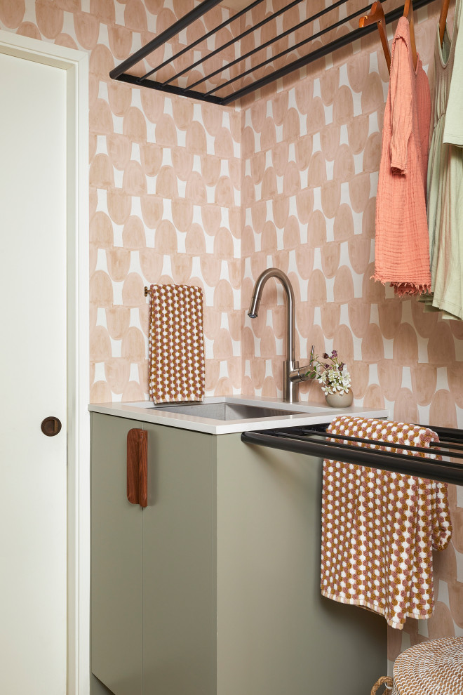 Retro inredning av en liten grå parallell grått tvättstuga enbart för tvätt, med en undermonterad diskho, släta luckor, gröna skåp, bänkskiva i kvarts, rosa väggar, betonggolv, en tvättmaskin och torktumlare bredvid varandra och brunt golv