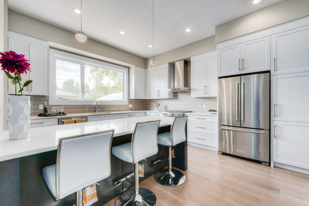 Photo of a large contemporary l-shaped separate kitchen in Calgary with an undermount sink, shaker cabinets, white cabinets, solid surface benchtops, beige splashback, porcelain splashback, stainless steel appliances, light hardwood floors, a peninsula and beige floor.