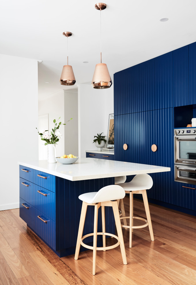 This is an example of a mid-sized contemporary galley eat-in kitchen in Melbourne with an undermount sink, blue cabinets, quartz benchtops, white splashback, engineered quartz splashback, stainless steel appliances, light hardwood floors, with island, brown floor and white benchtop.