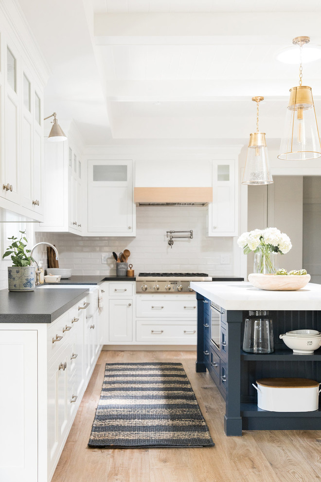 This is an example of a mid-sized beach style l-shaped kitchen pantry in Salt Lake City with a farmhouse sink, white cabinets, white splashback, light hardwood floors, with island and grey benchtop.