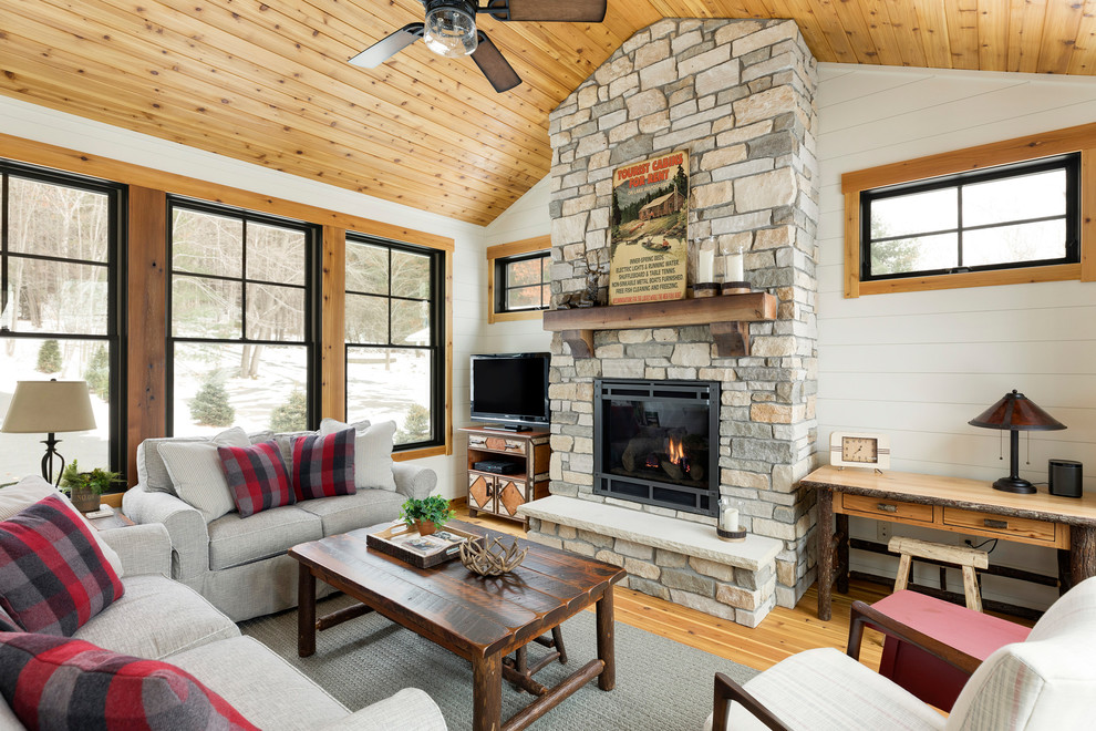 This is an example of a country family room in Minneapolis with white walls, light hardwood floors, a wood stove, a stone fireplace surround and a freestanding tv.