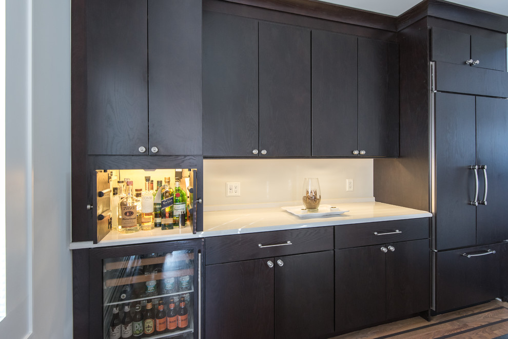 This is an example of a mid-sized transitional single-wall wet bar in Atlanta with no sink, flat-panel cabinets, dark wood cabinets, solid surface benchtops, white splashback and medium hardwood floors.