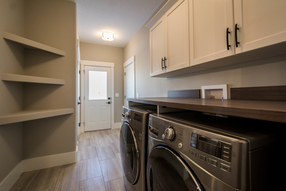Small transitional single-wall laundry cupboard in Calgary with shaker cabinets, white cabinets, solid surface benchtops, grey walls, porcelain floors, a side-by-side washer and dryer, beige floor and brown benchtop.