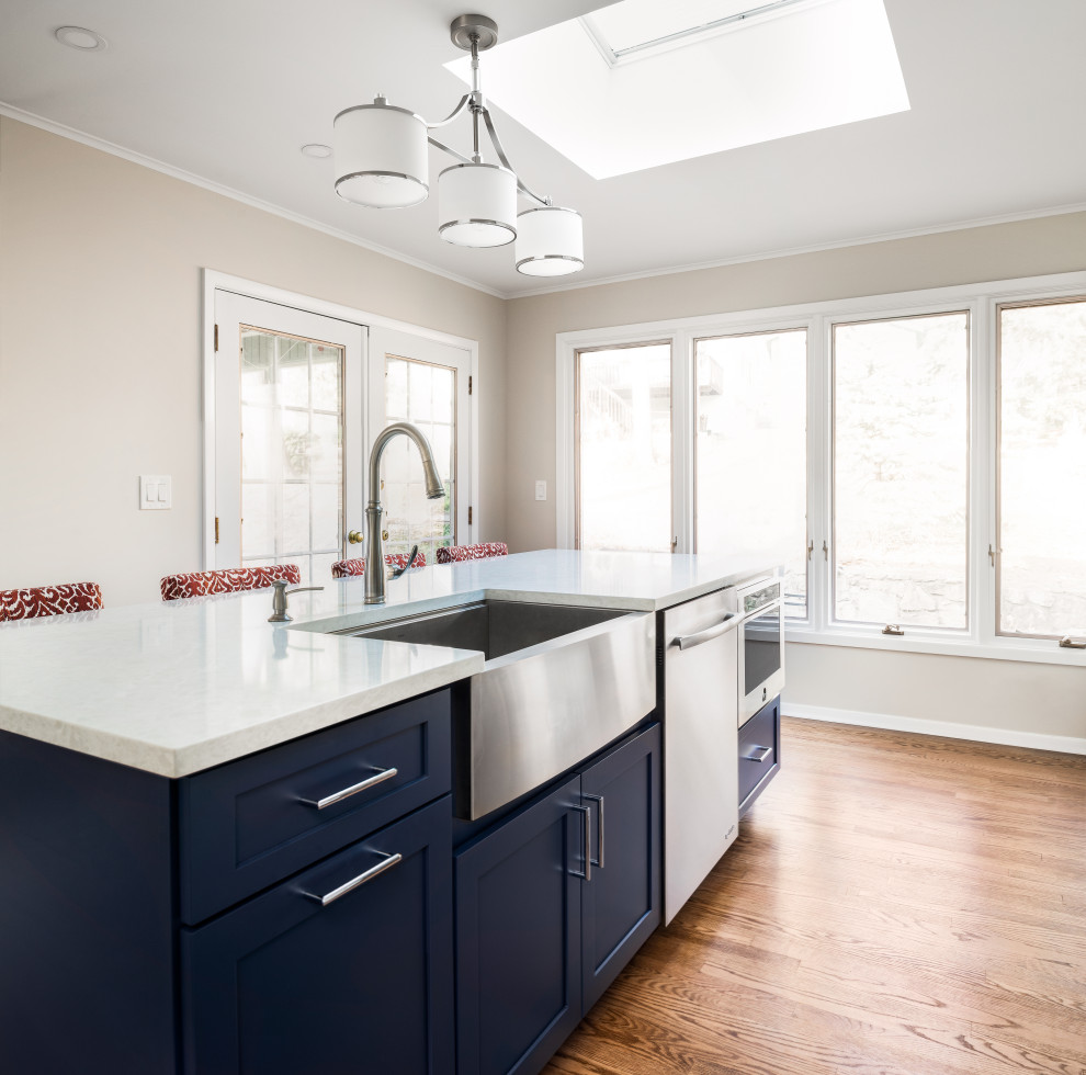 Example of a mid-sized transitional l-shaped medium tone wood floor eat-in kitchen design in New York with a farmhouse sink, shaker cabinets, white cabinets, quartz countertops, white backsplash, marble backsplash, stainless steel appliances, an island and white countertops