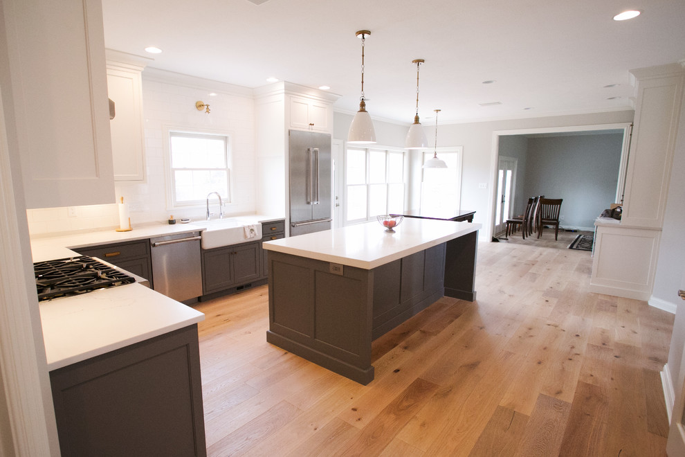 This is an example of a traditional eat-in kitchen in Indianapolis with a farmhouse sink, shaker cabinets, grey cabinets, white splashback, subway tile splashback, stainless steel appliances, medium hardwood floors, with island, brown floor and white benchtop.
