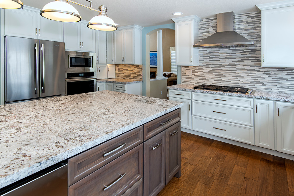 Example of a large transitional medium tone wood floor enclosed kitchen design with a farmhouse sink, recessed-panel cabinets, white cabinets, granite countertops, multicolored backsplash, matchstick tile backsplash, stainless steel appliances and an island