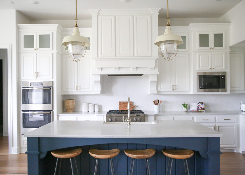 Inspiration for a large transitional u-shaped medium tone wood floor and brown floor eat-in kitchen remodel in Dallas with a farmhouse sink, raised-panel cabinets, white cabinets, quartz countertops, white backsplash, ceramic backsplash, stainless steel appliances, an island and white countertops