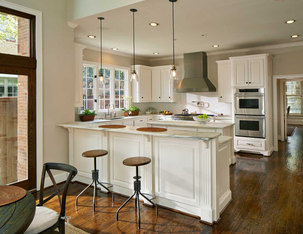 Example of a large classic u-shaped medium tone wood floor and brown floor open concept kitchen design in Dallas with an undermount sink, raised-panel cabinets, white cabinets, marble countertops, white backsplash, subway tile backsplash, stainless steel appliances, an island and white countertops