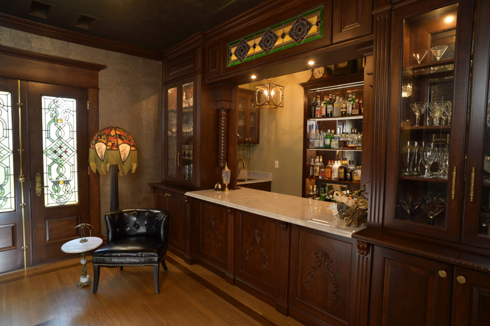 Inspiration for a mid-sized traditional single-wall wet bar in Other with a drop-in sink, medium wood cabinets, marble benchtops, medium hardwood floors, brown floor, brown benchtop and glass-front cabinets.