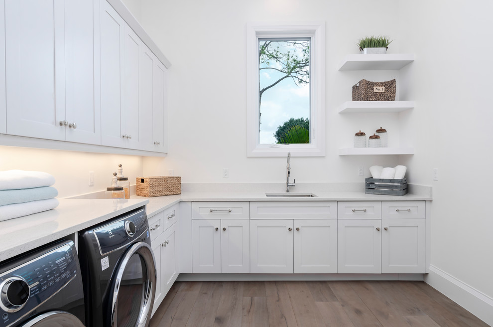 Inspiration for a large beach style l-shaped dedicated laundry room in Miami with a drop-in sink, shaker cabinets, white cabinets, quartzite benchtops, white walls, light hardwood floors, an integrated washer and dryer, brown floor and white benchtop.