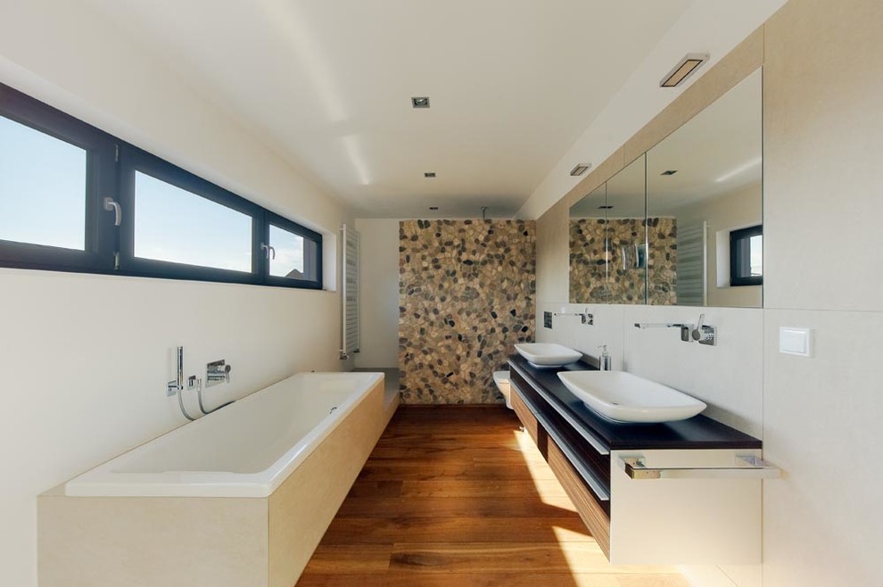 Inspiration for a large contemporary master bathroom in Cologne with a vessel sink, flat-panel cabinets, medium wood cabinets, a drop-in tub, an alcove shower, multi-coloured tile, pebble tile, white walls, medium hardwood floors and a wall-mount toilet.