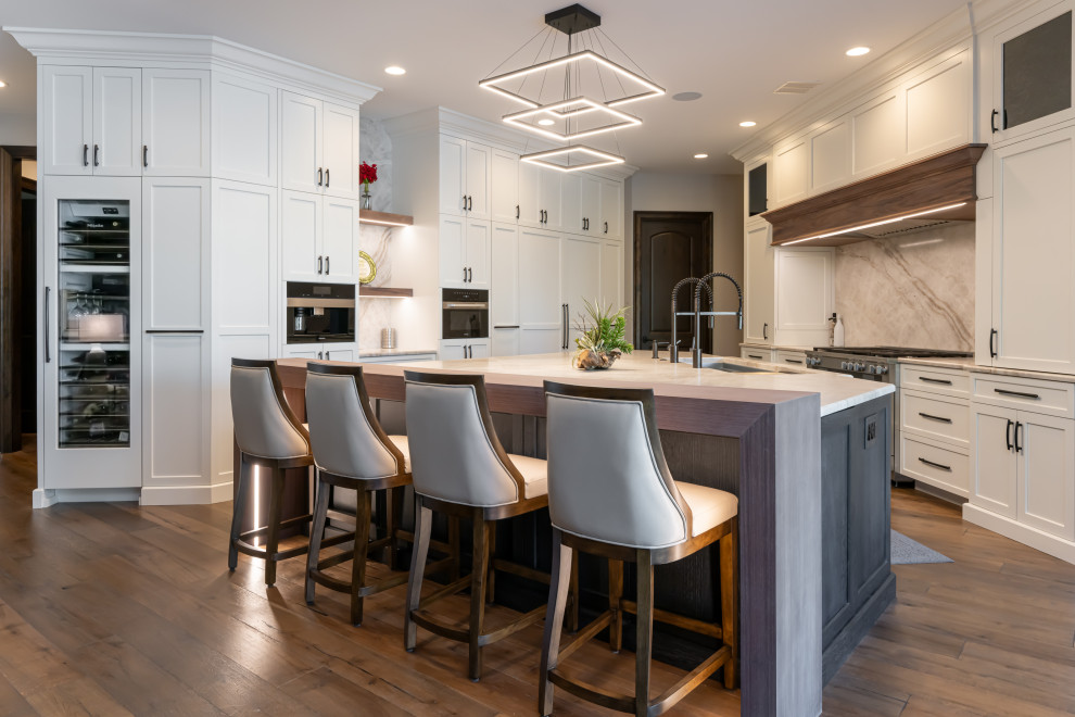 Large transitional medium tone wood floor and brown floor open concept kitchen photo with a double-bowl sink, flat-panel cabinets, white cabinets, quartzite countertops, beige backsplash, stainless steel appliances, an island and beige countertops