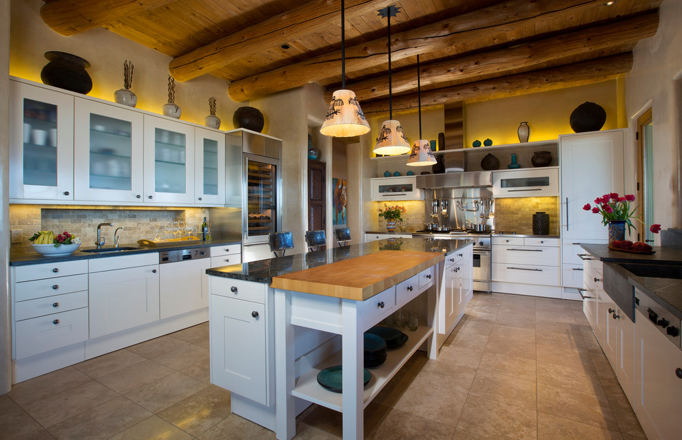 Large u-shaped kitchen in Albuquerque with a farmhouse sink, shaker cabinets, white cabinets, beige splashback, stainless steel appliances, with island, granite benchtops, stone tile splashback, ceramic floors and beige floor.