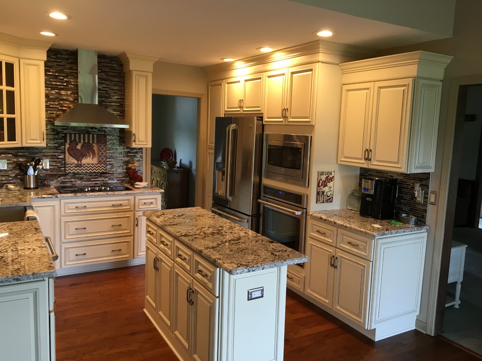 Mid-sized traditional u-shaped eat-in kitchen in Louisville with a farmhouse sink, raised-panel cabinets, white cabinets, granite benchtops, brown splashback, glass tile splashback, stainless steel appliances, medium hardwood floors, with island and brown floor.
