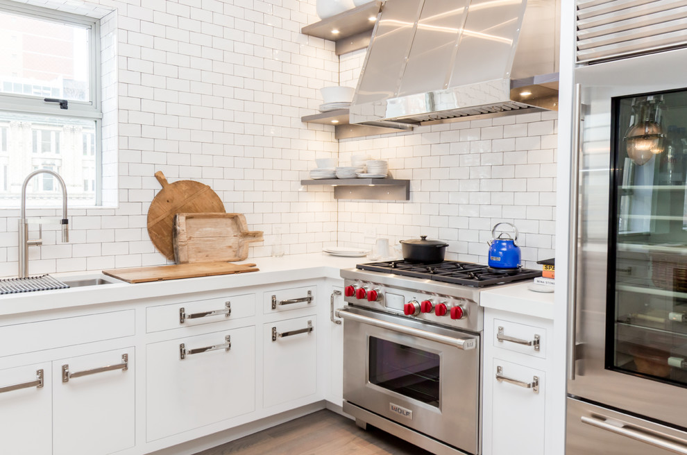 This is an example of a mid-sized industrial u-shaped kitchen in New York with white cabinets, quartz benchtops, white splashback, subway tile splashback, stainless steel appliances, light hardwood floors and a peninsula.