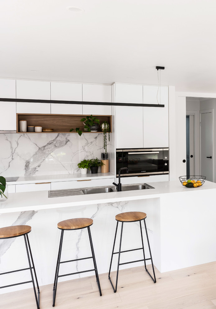 This is an example of a mid-sized modern galley open plan kitchen in Melbourne with a double-bowl sink, white cabinets, quartz benchtops, white splashback, marble splashback, white appliances, laminate floors, with island, brown floor and white benchtop.