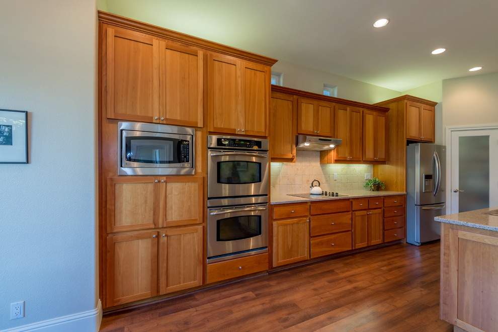 Photo of a large traditional l-shaped eat-in kitchen in Sacramento with an undermount sink, shaker cabinets, medium wood cabinets, granite benchtops, beige splashback, terra-cotta splashback, stainless steel appliances, vinyl floors, with island and brown floor.